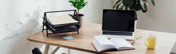 Notebook, laptop with blank screen and cup on working table, panoramic shot