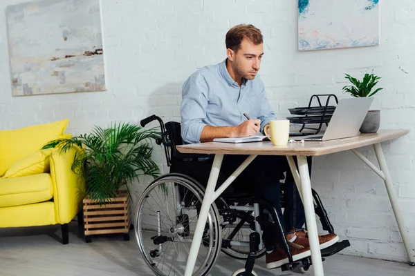 Geschäftsmann Rollstuhl Blickt Auf Laptop Und Schreibt Notizbuch Schreibtisch — Stockfoto