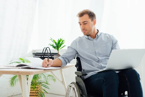 Geschäftsmann Rollstuhl Hält Laptop Und Schreibt Notizbuch Schreibtisch — Stockfoto