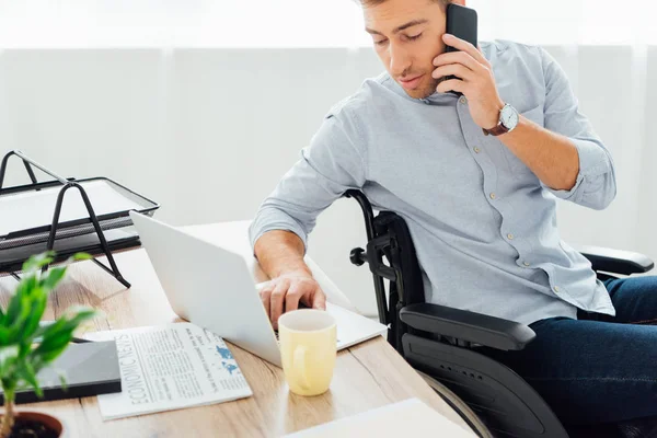Человек Инвалидной Коляске Разговаривает Смартфоне Использует Клавиатуру Ноутбука Столом — стоковое фото