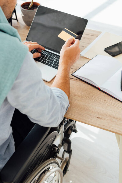 Cropped view of man in wheelchair holding credit card and using laptop with blank screen