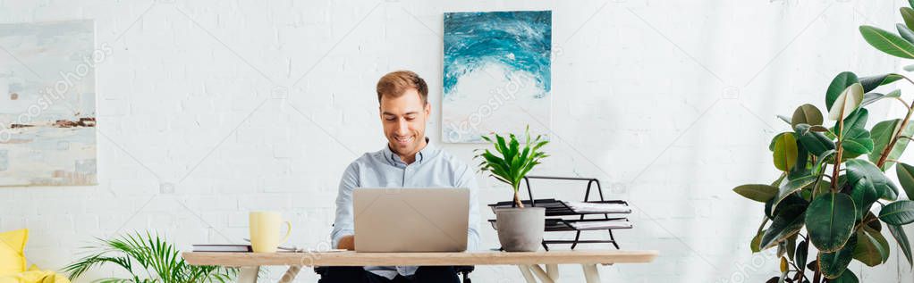 Smiling freelancer using laptop at desk in living room, panoramic shot