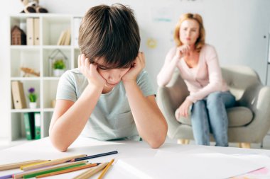 selective focus of sad kid with dyslexia and child psychologist on background  clipart