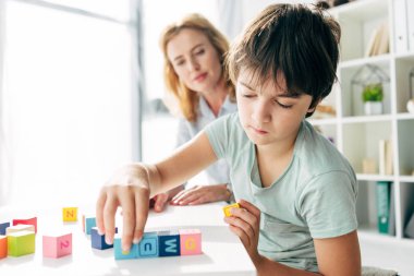 selective focus of kid with dyslexia playing with building blocks and child psychologist looking at it on background  clipart