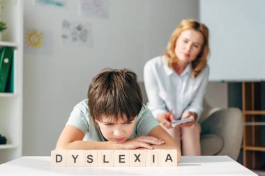 selective focus of sad kid with dyslexia sitting at table with wooden cubes with lettering dyslexia clipart
