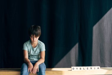 Disleksisi olan üzgün çocuk tahta küplerin yanında oturuyor ve siyah arkaplanda okuma güçlüğü çekiyor. 