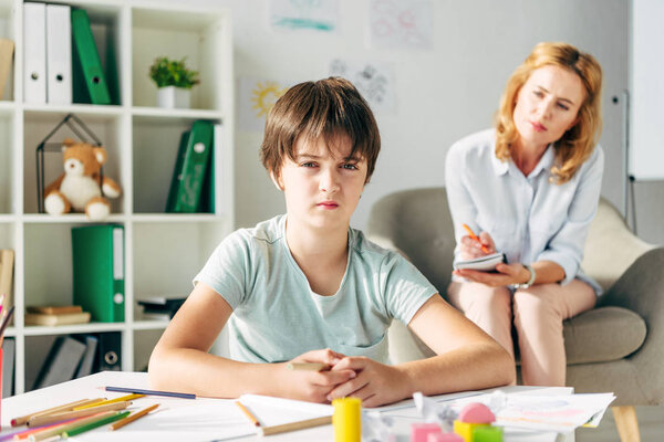 selective focus of kid with dyslexia looking at camera and child psychologist sitting on background 