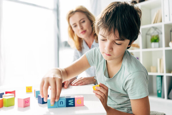 selective focus of kid with dyslexia playing with building blocks and child psychologist looking at it on background 