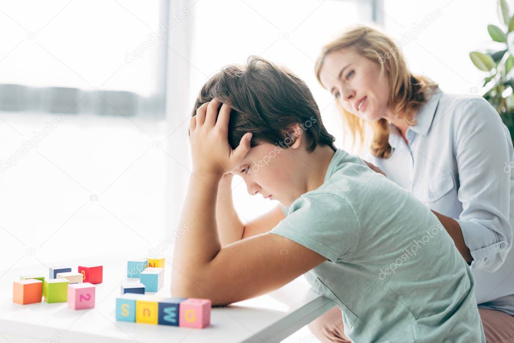 side view of sad kid with dyslexia looking at building blocks and child psychologist looking at him 