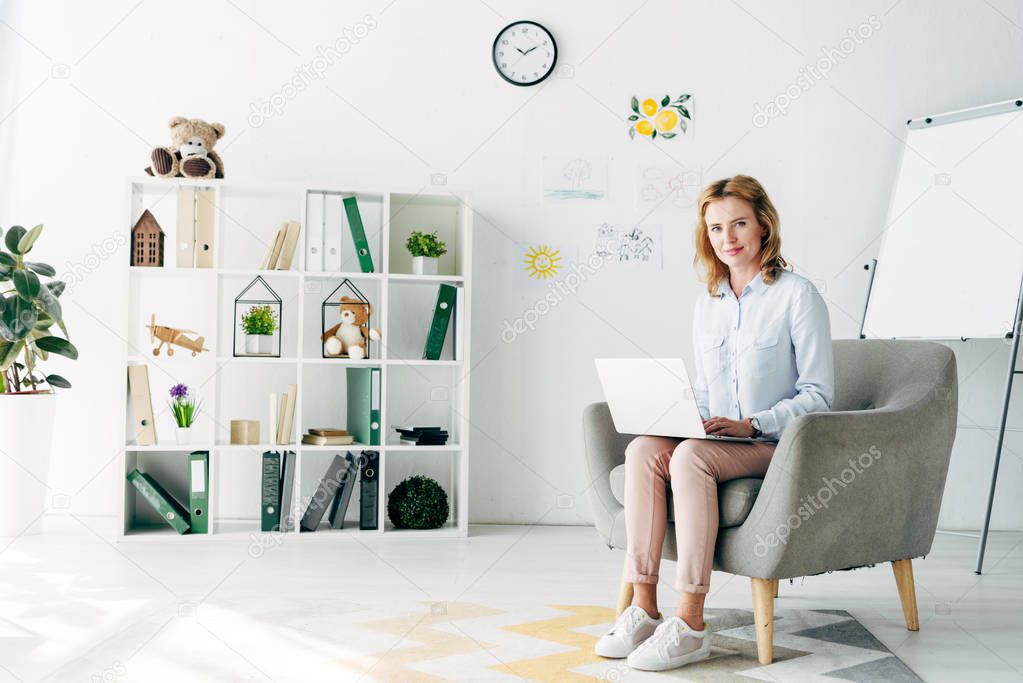 attractive child psychologist in shirt holding laptop and sitting on armchair 