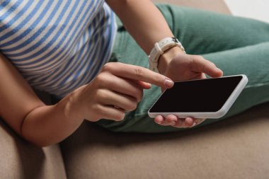 cropped view of woman using smartphone with blank screen while sitting on sofa clipart