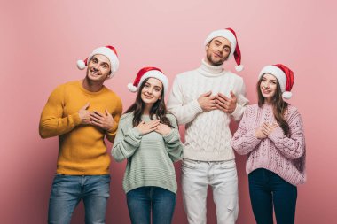 Süveter ve Noel Baba şapkalı mutlu arkadaşlar kalplerin üzerinde el ele tutuşuyorlar, pembe renkte izole olmuşlar.