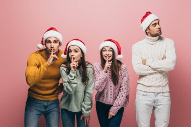 Süveter ve Noel Baba şapkalı neşeli arkadaşlar. Pembe renkte izole edilmiş sessizlik sembolleri gösteriyorlar.
