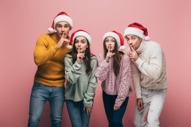 Süveter ve Noel Baba şapkalı mutlu arkadaşlar. Pembe renkte izole edilmiş sessizlik sembolleri gösteriyorlar.