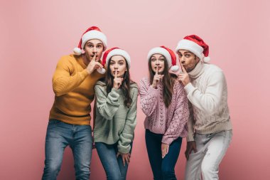 Süveter ve Noel Baba şapkalı mutlu arkadaşlar. Pembe renkte izole edilmiş sessizlik sembolleri gösteriyorlar.