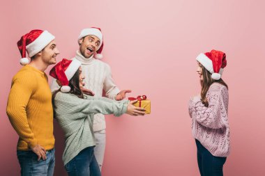 Noel Baba şapkalı mutlu arkadaşlar. Pembe renkte izole edilmiş kızlara Noel hediyesi veriyorlar.