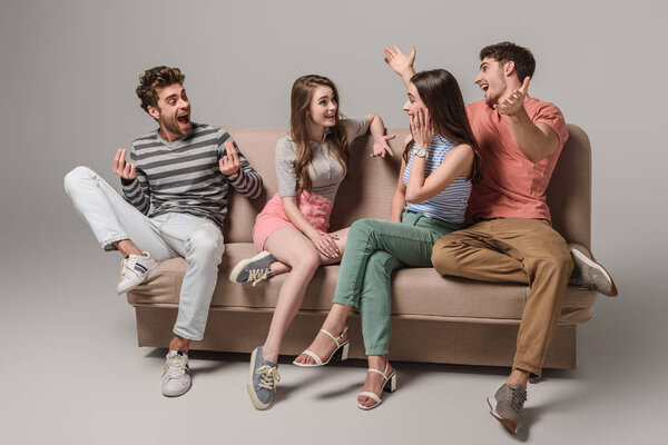 emotional young friends talking and sitting on sofa on grey 