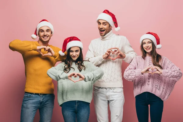 Süveter ve Noel Baba şapkalı mutlu arkadaşlar. Kalp sembolleri gösteriyorlar. Pembe renkte izole edilmiş.