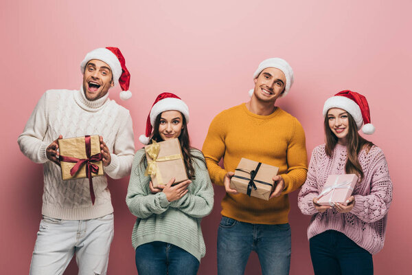 cheerful friends in santa hats holding christmas presents, isolated on pink