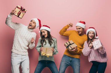 Noel Baba şapkalı heyecanlı arkadaşlar Noel hediyeleri taşıyorlar, pembe renkte izole edilmişler.