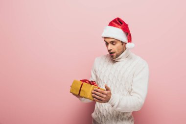 Şok olmuş süveter ve Noel Baba şapkalı bir adam. Elinde Noel hediyesi var. Pembe renkte izole edilmiş.