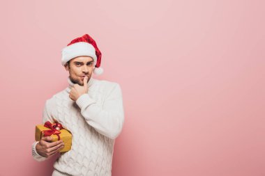 Süveter ve Noel Baba şapkalı düşünceli bir adam. Elinde Noel hediyesi var. Pembe renkte izole edilmiş.