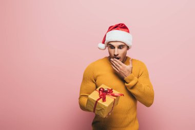 Şok olmuş süveter ve Noel Baba şapkalı adam pembelerle izole edilmiş Noel hediyesine bakıyor.