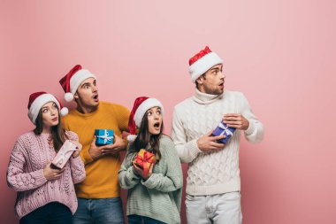 Noel Baba şapkalı şok olmuş arkadaşlar ellerinde hediyeler, pembe renkte izole edilmiş.