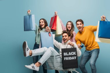 excited young friends having fun with shopping bags in shopping cart on black friday, isolated on blue clipart