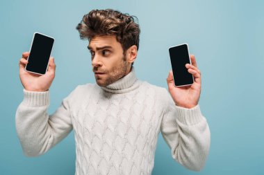 Duygusal adam boş ekranlı iki akıllı telefona bakıyor, mavi ekranda izole edilmiş.