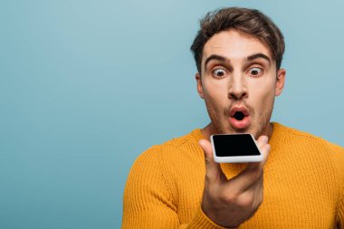 Şok olmuş yakışıklı adam akıllı telefon kullanıyor, mavi renkte izole edilmiş.