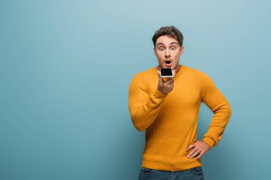 Şok olmuş yakışıklı adam akıllı telefon kullanıyor, mavi renkte izole edilmiş.