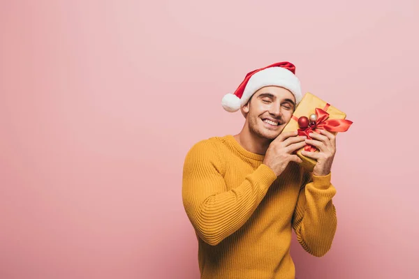 セーターを着た笑顔の男とクリスマスプレゼント箱を持ったサンタの帽子ピンクに隔離された — ストック写真