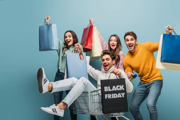 excited young friends having fun with shopping bags in shopping cart on black friday, isolated on blue