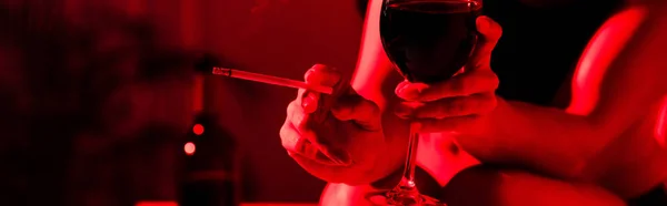 partial view of woman holding cigarette and glass of wine in red light, panoramic shot