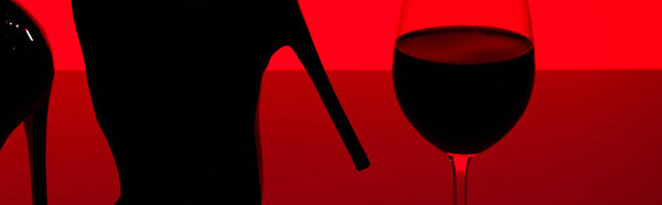 cropped view of silhouette of female feet in heels with glass of wine isolated on red, panoramic shot