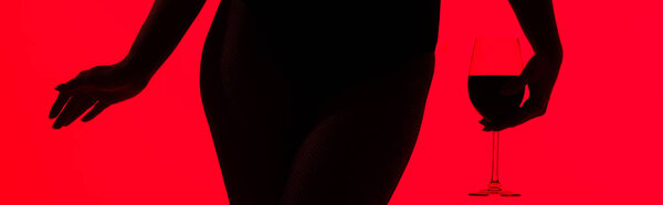 cropped view of black silhouette of sexy girl holding glass of wine isolated on red, panoramic shot