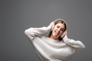 attractive funny girl in white sweater and earmuffs sticking tongue out, isolated on grey clipart
