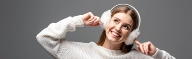 panoramic shot of attractive happy girl posing in white sweater and earmuffs, isolated on grey clipart