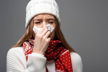 ill woman in knitted hat having runny nose and holding paper napkin, isolated on grey clipart