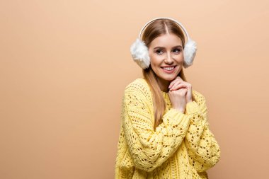 Sarı süveter ve kulak ısıtıcı giymiş, bej rengi elbiseler içinde güzel gülümseyen bir kadın.