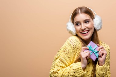 Sarı kazaklı ve kulaklıklı neşeli bir kadın Noel hediyesi taşıyor, bej renkte izole edilmiş.