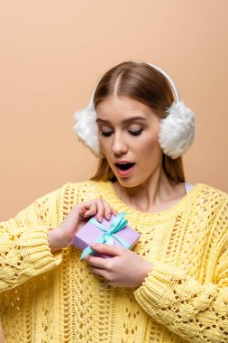 Sarı kazaklı ve kulak ısıtıcılarıyla sürpriz bir kadın Noel hediyesi taşıyor, bej rengi bir elbiseyle izole edilmiş.