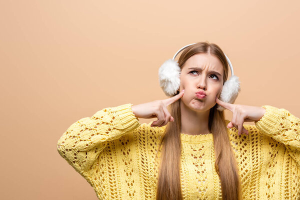 beautiful disappointed woman in sweater and earmuffs touching her cheeks, isolated on beige