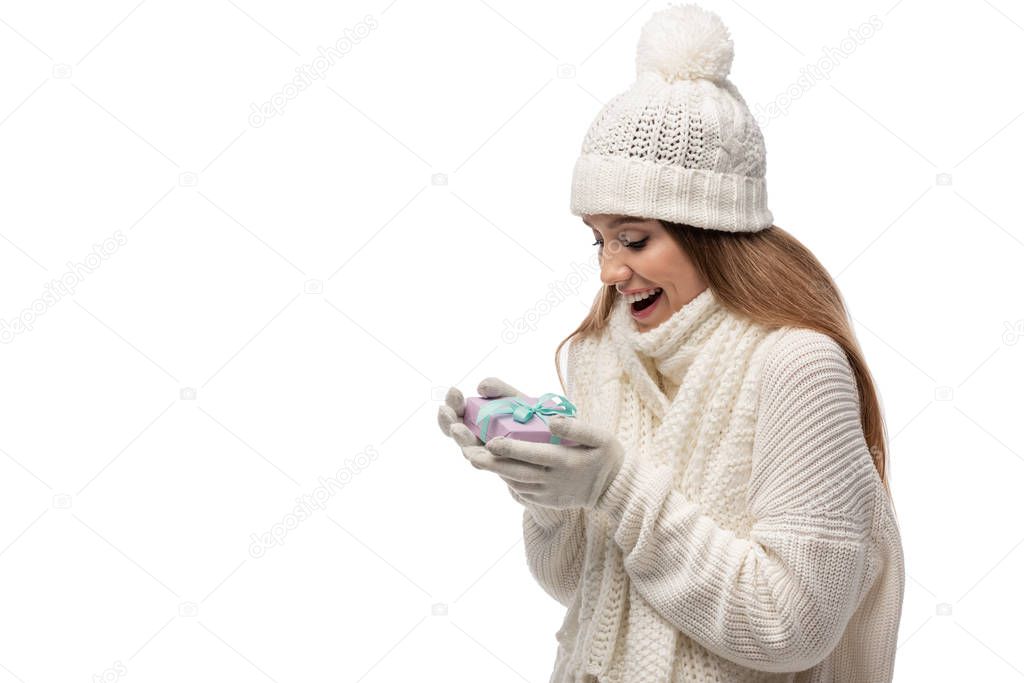 excited woman in scarf, gloves and hat looking at gift box, isolated on white