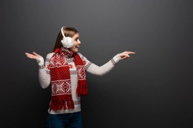 Noel süveteri, atkısı ve kulaklığı olan heyecanlı kadın gri bir şeyi işaret ediyor.