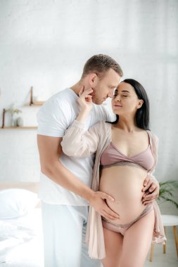 Yakışıklı koca yatak odasında gülümseyen hamile karısına sarılıyor. 