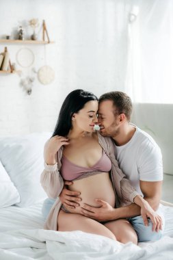 Mutlu koca yatakta çekici hamile karısına sarılıyor.