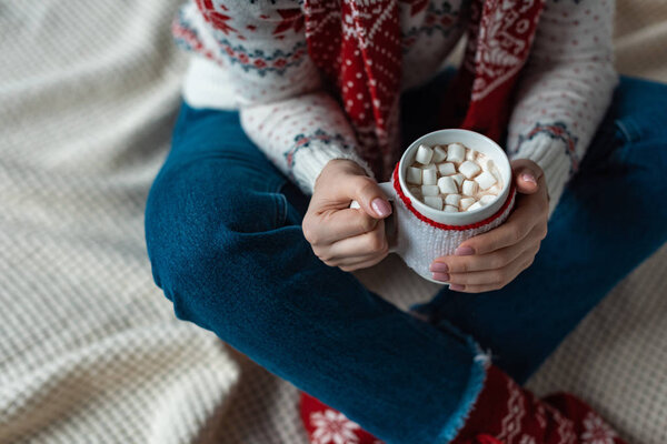 cropped view of woman in warm sweater holding cup of cocoa with marshmallow and sitting on blanket
