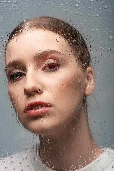 attractive sad woman on grey, beyond wet glass with drops 